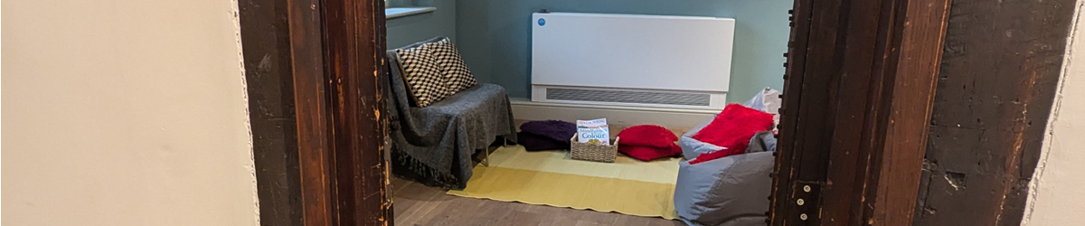 The National Civil War Centre's Quiet Space looking in from the Tudor Hall doorway. With the timber door framing it on either, side, you can see a yellow rug in between a grey chair and two bean bags. There are fluffy cushions on all the seats and on the floor which also has a basket of colouring books and coloured pencils.